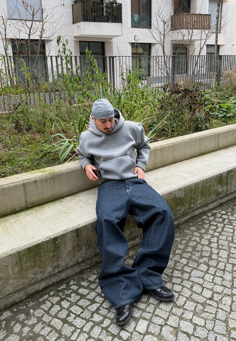 Homme en sweat à capuche gris et casquette assis sur un muret en béton près de la verdure et d'un trottoir pavé, tenant un smartphone.