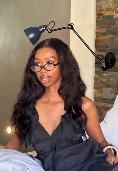 Top negro de satén con volantes y un escote profundo. Cabello largo y ondulado. Gafas redondas. Una lámpara azul en ángulo está montada en la pared de fondo.