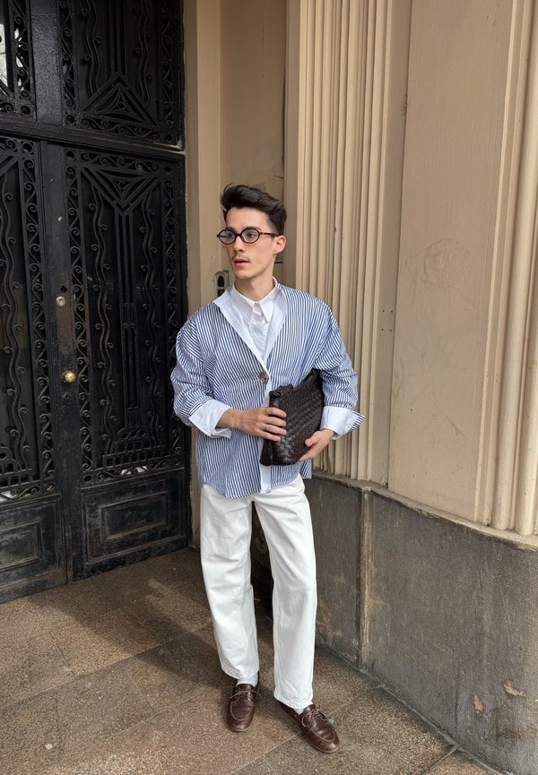 Jonge man met bril die een gestreept blauw-wit hemd, witte broek, bruine schoenen draagt en een zwarte geweven clutch vasthoudt bij een gebouwingang.