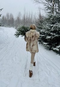 Personne portant un manteau de fourrure et des bottes marche sur un chemin enneigé en portant un sapin, devant un paysage de arbres couverts de neige.