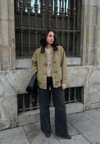 Chaqueta de algodón color caqui con un cuello marrón, suéter ligero y pantalones vaqueros de pierna ancha negros. Un bolso negro texturizado complementa el atuendo.