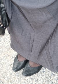 Bailarinas de cuero negro con puntas afiladas, usadas sobre medias gris con pequeños lunares, junto a una falda gris fluida sobre una superficie de grava.