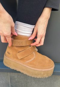 Tan nubukist kinga Velcro rihmaga ja "UGG" logoga, paksu kummist tallaga ja valge sisetallaga, halli tausta peal.
