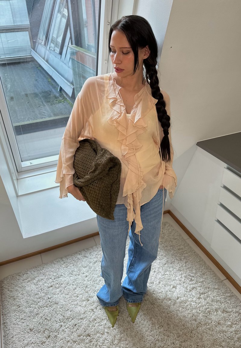 Blouse froncée rose clair, jeans bleus taille haute, chaussures vertes à bout pointu, tenant un sac olive texturé, debout à l'intérieur près d'une fenêtre.