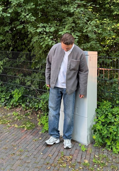 Chaqueta de forro polar gris con un bolsillo frontal, camisa blanca debajo, jeans azules y zapatillas grises, con un fondo de vegetación y una caja de servicios.