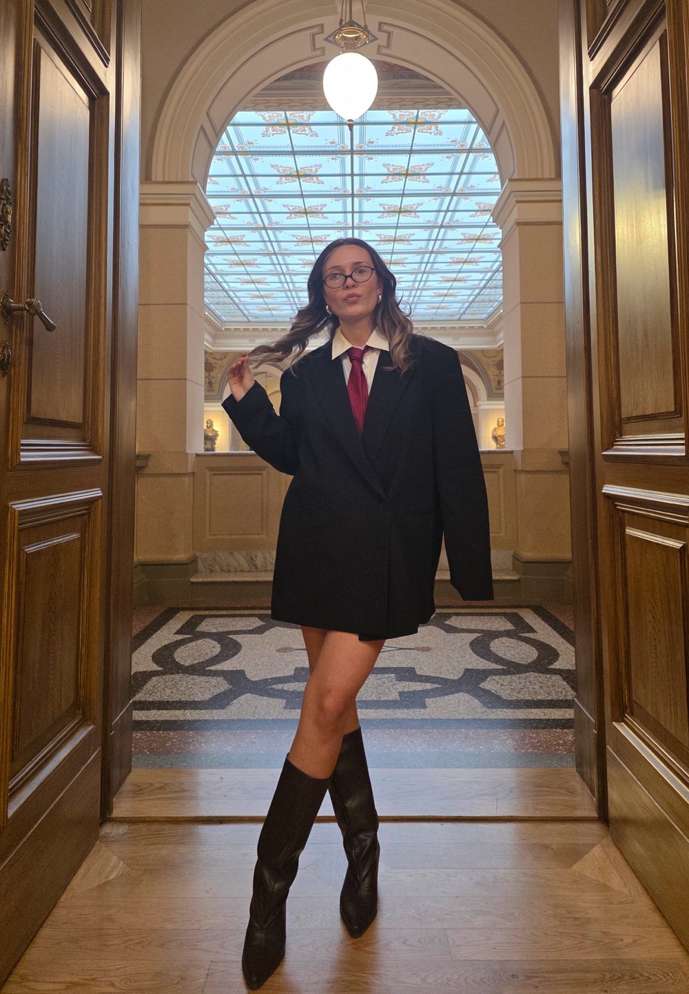 Obszerna czarna marynarka z białą koszulą i czerwonym krawatem, zestawiona z czarnymi kozakami sięgającymi kolan. W tle ozdobny łuk i wzorzysta podłoga.