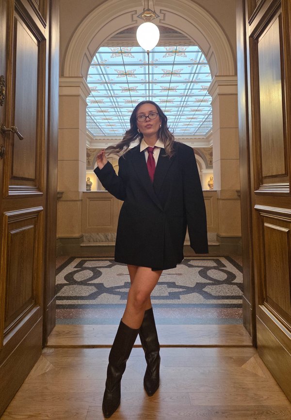 Blazer nero oversize con camicia bianca e cravatta rossa, abbinati a stivali neri al ginocchio. Sullo sfondo un arco decorato e pavimentazione con motivi.