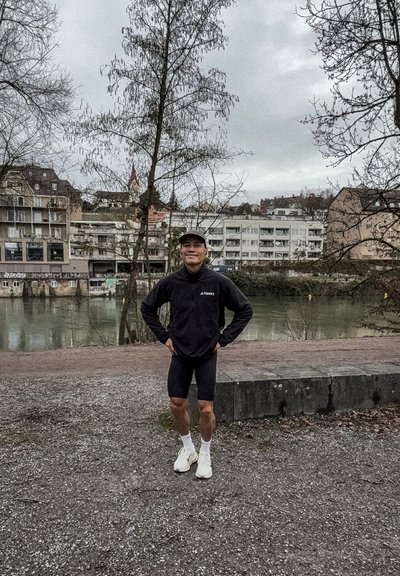Joven con ropa deportiva de pie con las manos en las caderas cerca de un río, con árboles sin hojas y edificios urbanos de fondo en un día nublado.