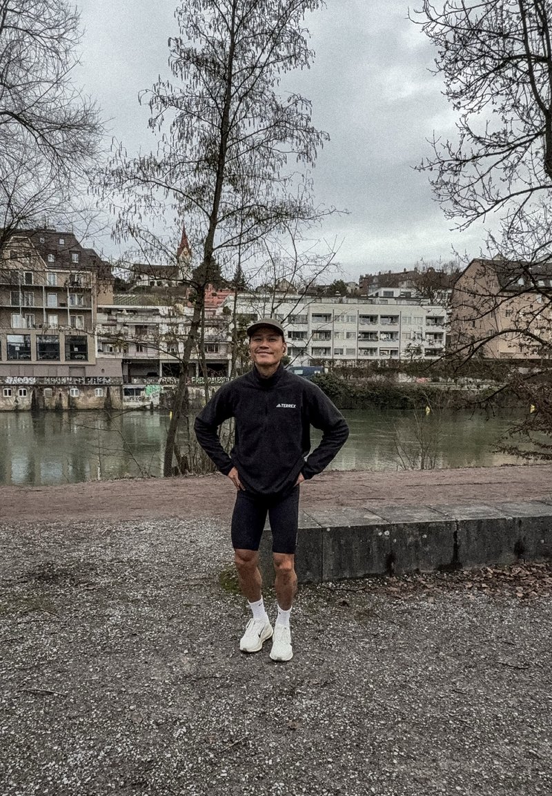 Giovane uomo in abbigliamento sportivo in piedi con le mani sui fianchi vicino a un fiume, con alberi spogli e edifici urbani sullo sfondo in una giornata nuvolosa.