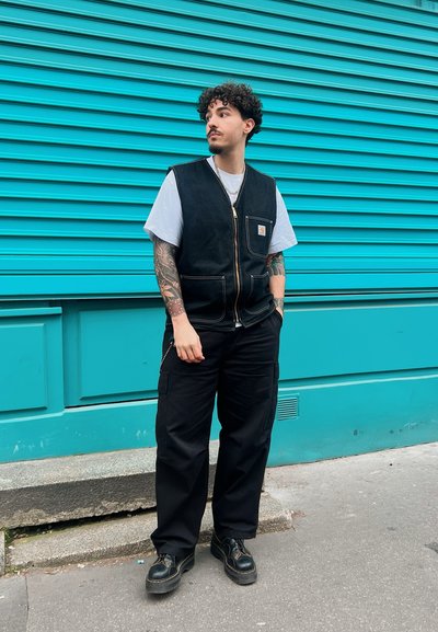 Chaleco de mezclilla negro con cremallera frontal y bolsillo con parche, usado sobre una camiseta blanca. Combinado con pantalones cargo negros y zapatos negros de cordones.