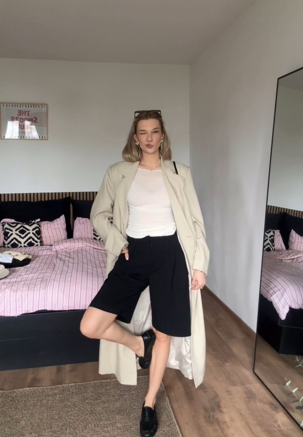 Mujer de pie en el dormitorio con gabardina beige, top blanco, pantalones cortos negros y zapatos negros, posando con una pierna levantada y la mano en el bolsillo.