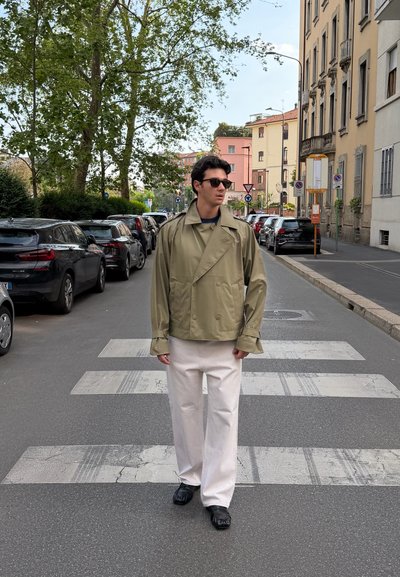 Joven con gafas de sol, chaqueta beige y pantalones blancos caminando por un paso de peatones de la ciudad con coches aparcados y edificios al fondo.