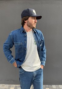 Blaue Jeans-Hemden mit Knöpfen, zwei Vordertaschen, graues T-Shirt mit Grafik darunter und blaue Jeans. Schwarze Mütze mit Logo vorne.