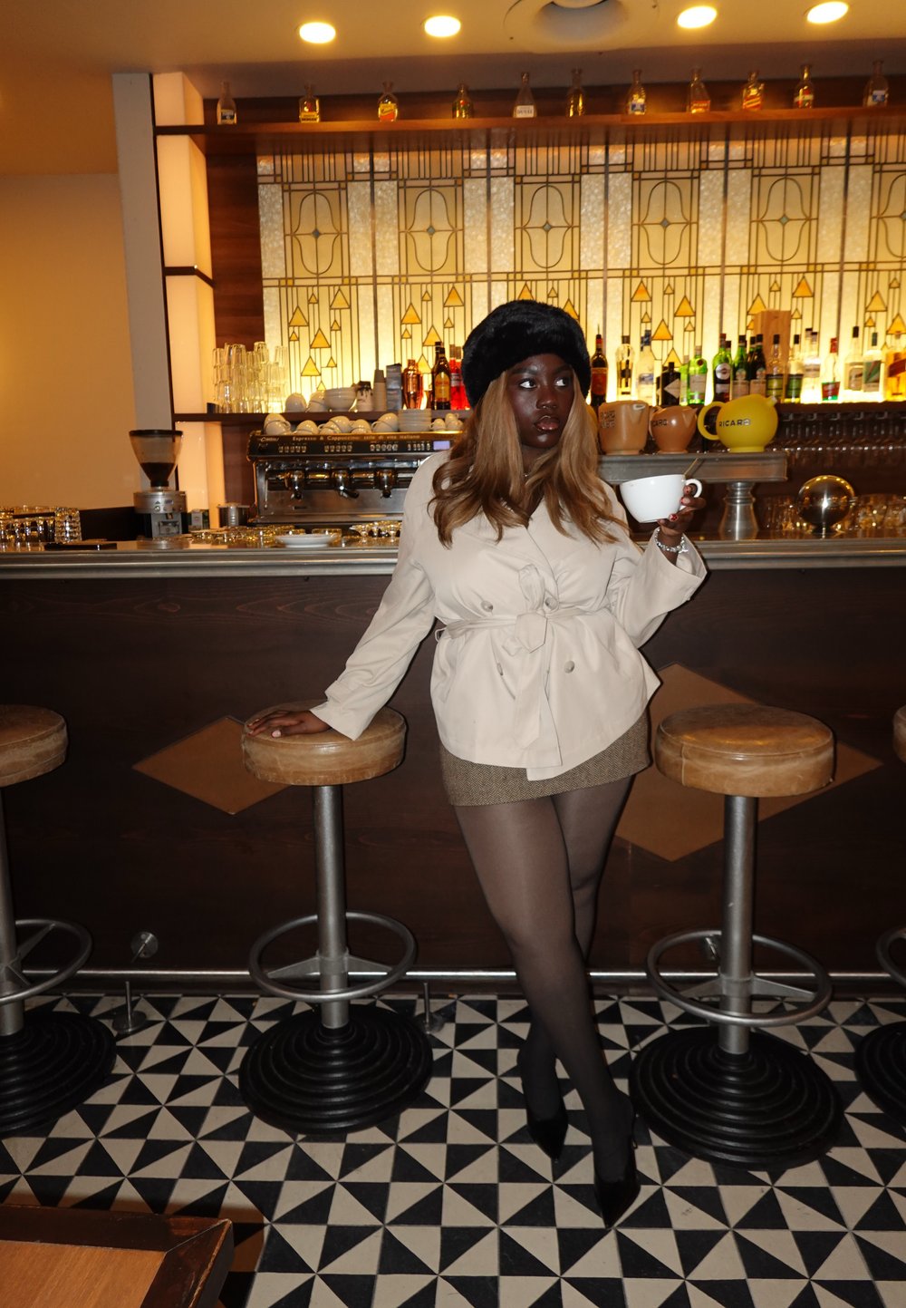 Beige trenchcoat med bånd, brun tweed mininederdel, sorte strømpebukser og højhælede sko. Holder en hvid kop foran en bar med geometriske fliser.