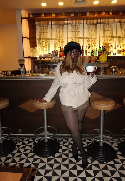 Abrigo trench beige con cinturón, minifalda de tweed marrón, medias negras y tacones altos. Sosteniendo una taza blanca, frente a un bar con azulejos geométricos.