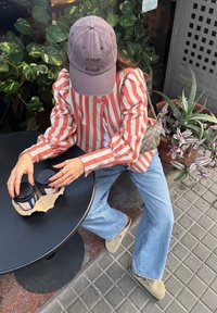 Stribet rød og hvid bluse med puffedeærmer, lyseblå flagrende jeans, grå kasket, holder to kaffekopper på et sort bord, omgivet af planter.