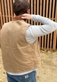 Béžová bavlnená vesta s hnedým kordovým golierom, bez rukávov, s jemnými textúrami, nosená cez sivú dlhú košeľu s rukávmi.