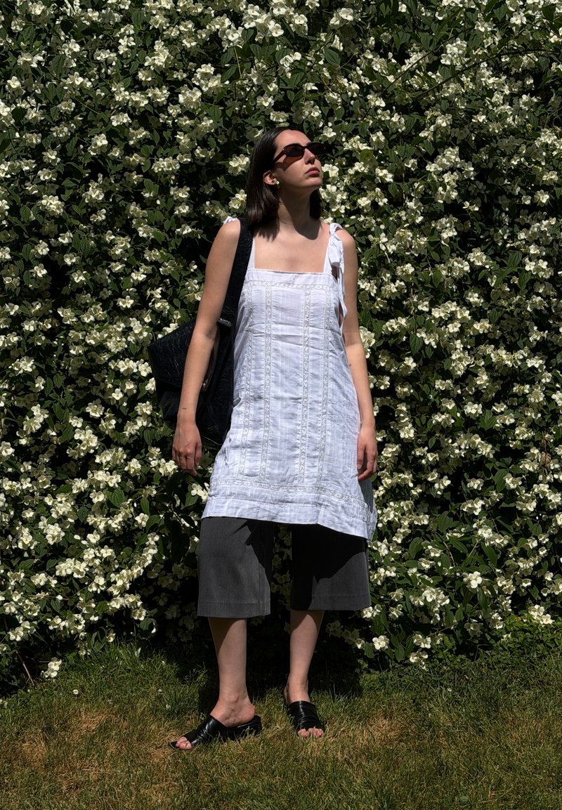 Femme portant une robe blanche et des lunettes de soleil foncées, debout sur l'herbe devant un buisson dense couvert de petites fleurs blanches.