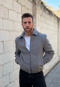 Άνδρας με κοντά καστανά μαλλιά και γένια, φοράει γκρι ζακέτα με φερμουάρ και λευκό πουκάμισο, στέκεται μπροστά σε ανοιχτόχρωμο πέτρινο τοίχο σε εξωτερικό χώρο.