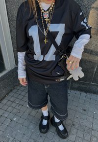 Fekete NFL mez ezüst 17-es számmal, egy fehér hosszú ujjú póló felett viselve. Sötét farmernadrág, fehér zoknik és vastag fekete cipők. Kiegészítők között rétegezett aranyláncok különböző medálokkal és egy kicsi plüss kulcstartó található.