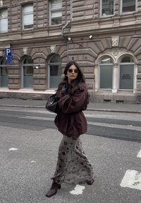 Giacca marrone oversize, guanti neri, gonna lunga a fiori in grigio chiaro e stivali marroni con tacco. Accessori includono occhiali da sole e borsa.