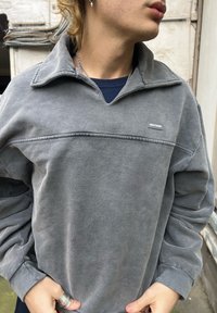 Šedý fleecový pulovr s polovičním zipem na límci, žebrovanými manžetami a malým logem na hrudi. Měkká textura s volným střihem.