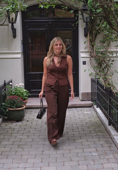 Blusa sin mangas marrón con un frente abotonado y pantalones de pierna ancha. Una bufanda marrón con estampado está atada al cuello. Bolso negro y sandalias.