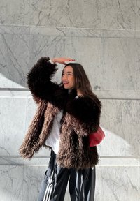 Jeune femme portant un manteau sombre et duveteux ainsi qu'un pantalon noir, souriant tout en se protégeant les yeux de la lumière du soleil devant un mur en marbre.