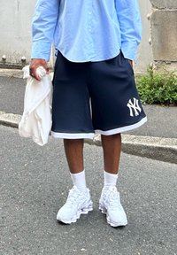 Ναυτικές βερμούδες με λευκή λεπτομέρεια και λογότυπο των New York Yankees, συνδυασμένες με λευκά πάνινα παπούτσια και κάλτσες μέχρι τον αστράγαλο. Ανοιχτό μπλε πουκάμισο με κουμπιά κρατιέται στο χέρι.