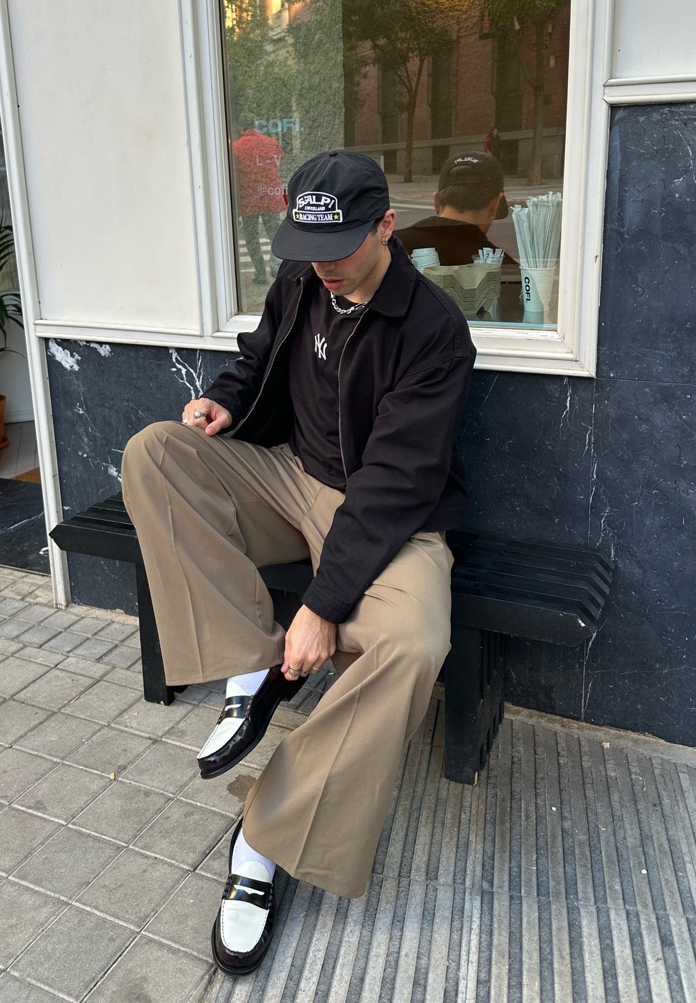 Sort jakke, sort kasket med logo, sorte og hvide loafers, beige bukser med vide ben, hvide sokker. Sidder på en sort bænk foran en marmorvæg.
