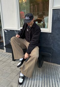 Sort jakke, sort kasket med logo, sorte og hvide loafers, beige bukser med vide ben, hvide sokker. Sidder på en sort bænk foran en marmorvæg.