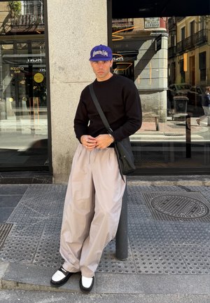 Hombre con suéter negro, pantalones anchos beige, zapatos blancos y negros y gorra azul parado en la acera de la ciudad cerca de una ventana del edificio.