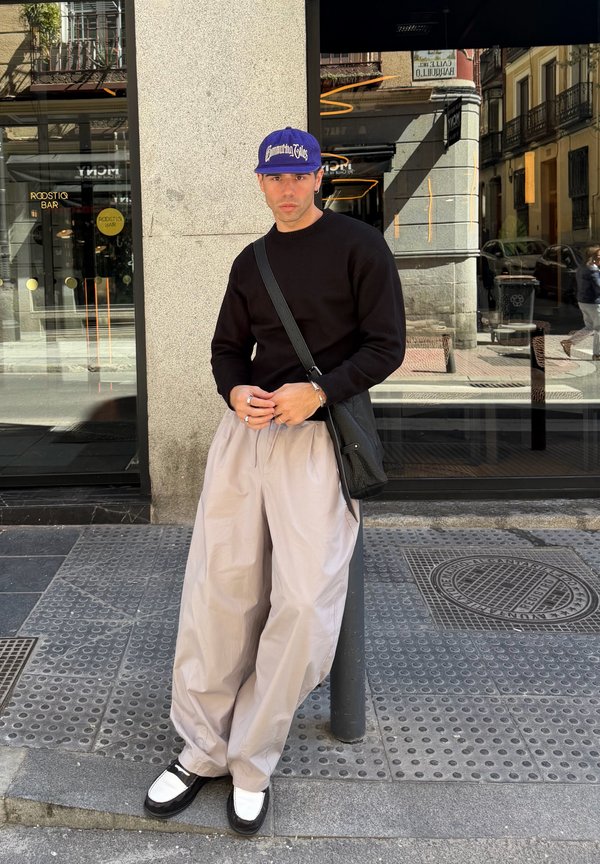 Homme portant un pull noir, un pantalon large beige, des chaussures blanches et noires, et une casquette bleue, debout sur un trottoir en ville près d'une fenêtre de bâtiment.