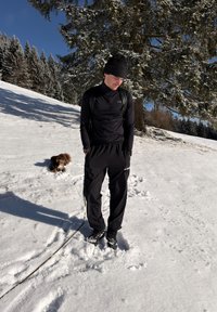 Homme en tenue d'hiver noire et chapeau debout sur la neige avec un petit chien brun à proximité sous un arbre couvert de neige par une journée claire.