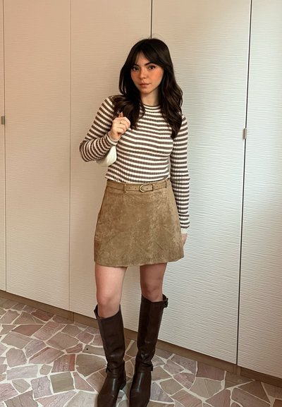 Top de manga larga a rayas en marrón y blanco, combinado con una mini falda de ante en tono arena. Botas marrones hasta la rodilla completan el conjunto. Bolso blanco en la mano.