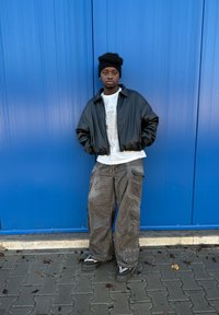 Fekete bomberdzseki fehér grafikás póló felett, szürke túlságosan bő cargo nadrággal és fekete cipővel párosítva. Kék fal a háttérben.