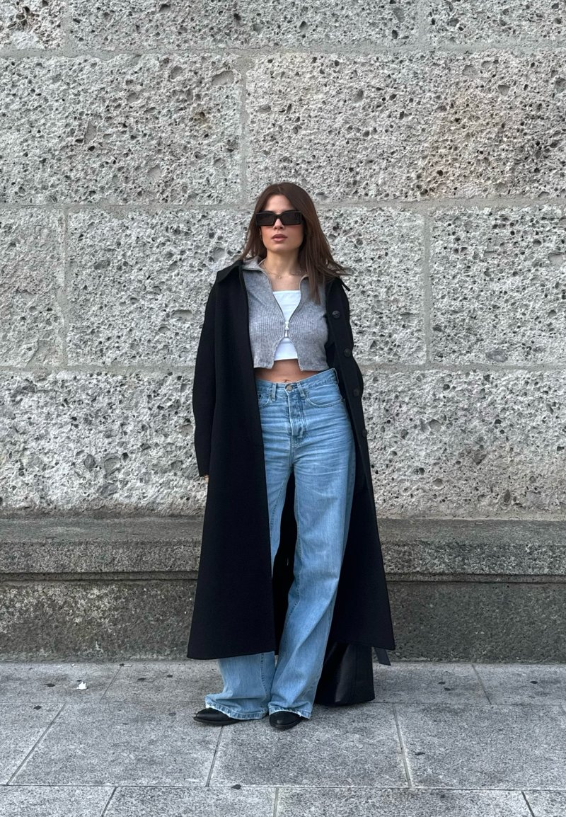 Donna con cappotto lungo nero, maglione grigio corto con zip, top bianco, jeans blu a gamba larga, scarpe nere e occhiali da sole scuri, in piedi contro un muro di pietra.