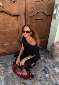 Svart ribbet off-shoulder topp, svarte leggings, bordeaux håndbag med krokodillestruktur og dusk, sitter på brostein nær en tre dør.