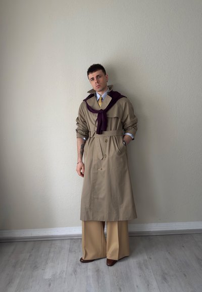 Joven con gabardina beige, pantalones de pierna ancha y zapatos marrones, de pie contra una pared lisa con un suéter morado sobre los hombros.