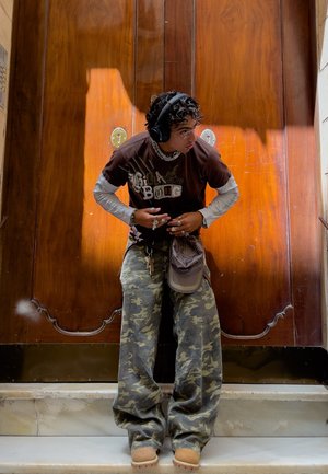 Camiseta gráfica marrón superpuesta a una camiseta de manga larga, pantalones de camuflaje de corte holgado y zapatos marrones. Los accesorios incluyen auriculares y anillos.