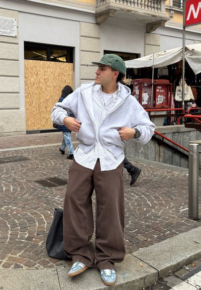 Sudadera gris con cremallera y corte holgado, camiseta a rayas blanca debajo, pantalones anchos marrones y zapatillas azul claro con suela beige.