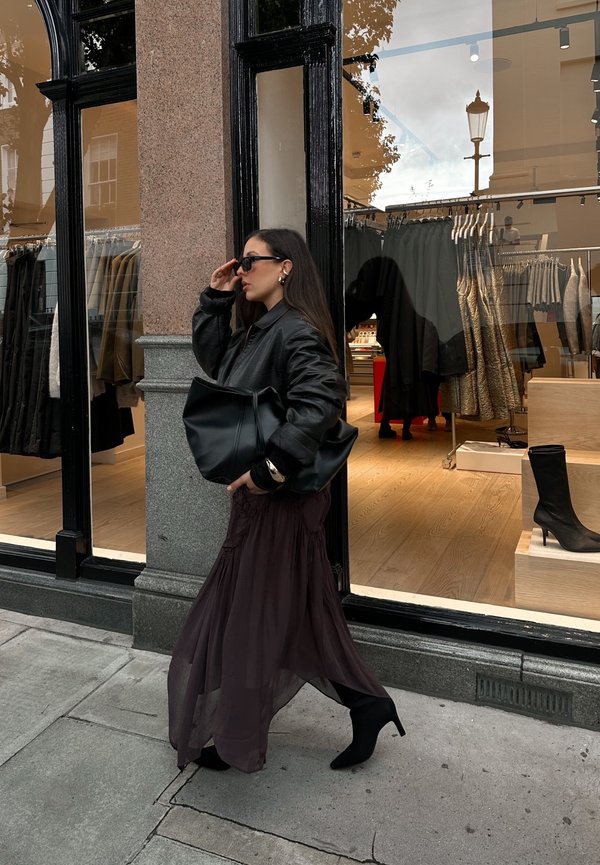 Veste en cuir noir oversize, jupe maxi transparente violet foncé, bottines noires, grand sac noir et lunettes de soleil. Vitrine de magasin en arrière-plan.
