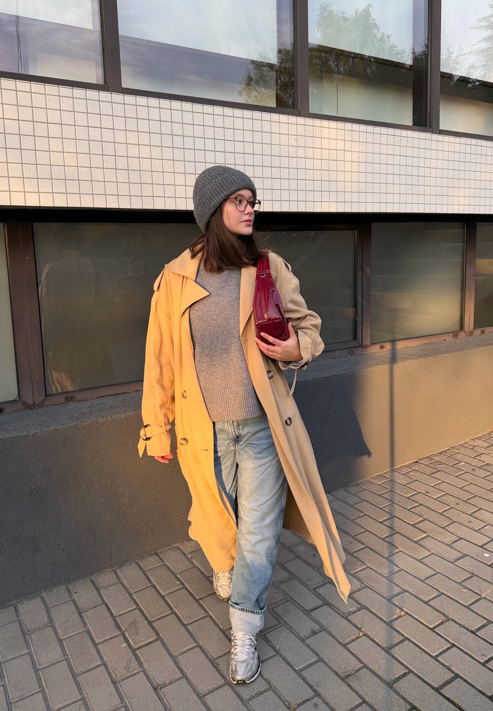 Trenci bej, pulover gri, blugi albaștri. Căciulă tricotată gri, geantă roșie, adidași argintii. Mergând pe pavajul de cărămidă lângă ferestre mari.