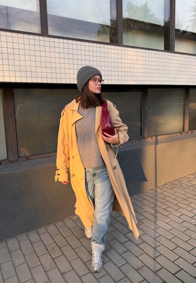 Abrigo trench beige, suéter gris, jeans azules. Gorro tejido gris, bolso rojo, zapatillas plateadas. Caminando sobre pavimento de ladrillo cerca de grandes ventanas.