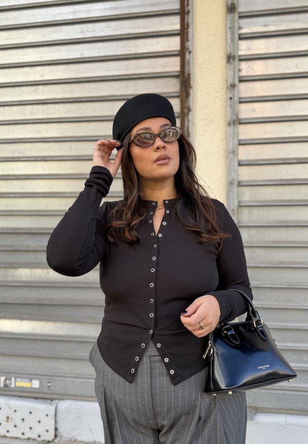 Vrouw met zwarte hoed en bril, zwarte top met knopen, grijze broek met krijtstreep, houdt een zwarte handtas vast, staat voor een metalen rolluik.