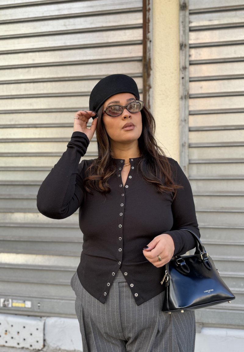 Frau mit schwarzem Hut und Brille, schwarzem Knopfoberteil, grauer Nadelstreifenhose, hält eine schwarze Handtasche, steht vor einem Metallrollladen.