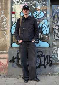 Giacca nera in nylon con chiusure a scatto, jeans grigi oversize, scarpe nere e un cappello nero con logo bianco, in piedi davanti a un muro con graffiti.
