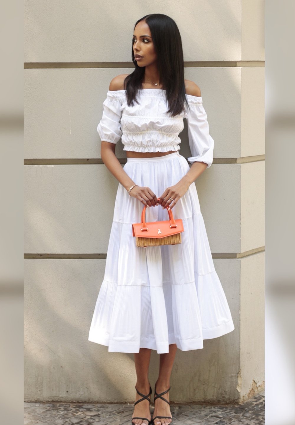 Weiße schulterfreie Bluse mit Rüschendetails, kombiniert mit einem fließenden weißen Stufenkleid. Hält eine kleine orangefarbene und geflochtene Handtasche. Schwarze Riemchen-Heels.