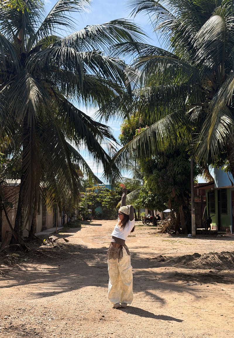 Femme portant un pantalon ample et un foulard sur la tête s'étire les bras levés sous de grands palmiers sur un chemin de terre ensoleillé.