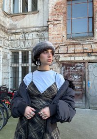 En person bærer en sort dunjakke over en hvid skjorte og en brun ternet slipkjole. Tilbehør inkluderer store sølvøreringe og ringe.